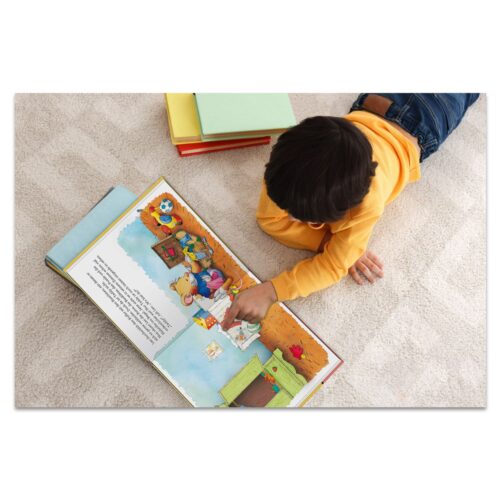 Kind im gelben Pullover liest ein buntes Geschichtenbuch auf einem Teppich, daneben stehen gestapelte Bücher.